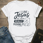 A white short-sleeve graphic T-shirt laid flat on a rustic wooden surface, styled with blue jeans and a Bible. The shirt features black text reading 'I LOVE Jesus BUT YOU'RE REALLY PUSHING IT!' alongside a cartoon bear illustration.