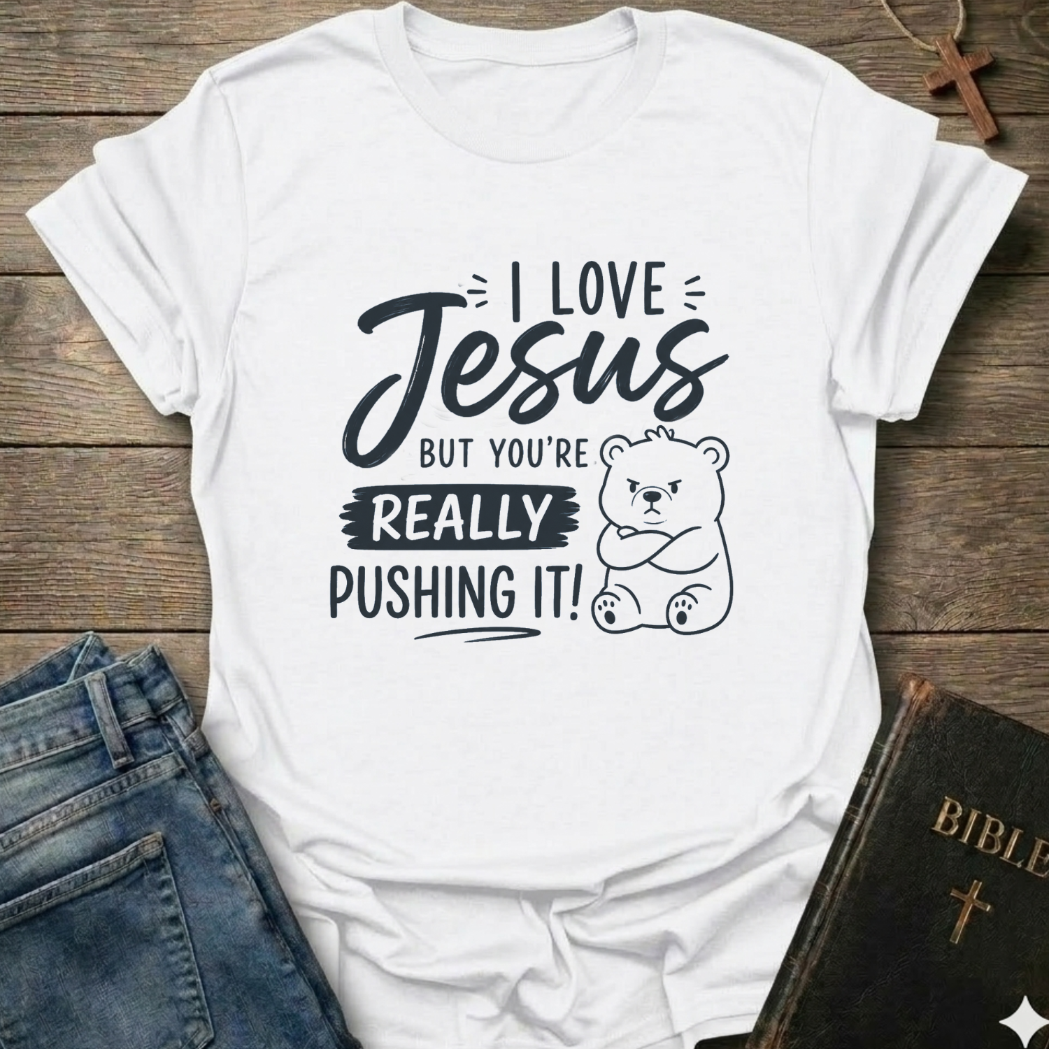A white short-sleeve graphic T-shirt laid flat on a rustic wooden surface, styled with blue jeans and a Bible. The shirt features black text reading 'I LOVE Jesus BUT YOU'RE REALLY PUSHING IT!' alongside a cartoon bear illustration.