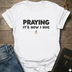White t-shirt with 'PRAYING IT'S HOW I HUG' text Christian faith apparel relatable scripture tee conversation starter on a wooden surface with jeans and a Bible.