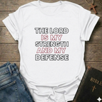 White t-shirt with 'The Lord is my strength and my defense' Exodus 15:2 bold faith apparel Faith Forward on a wooden surface with jeans and a Bible.