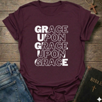 Maroon t-shirt with 'GRACE UPON GRACE' Christian T-Shirt John 1:16 bold faith apparel text on a wooden surface with jeans and a Bible.