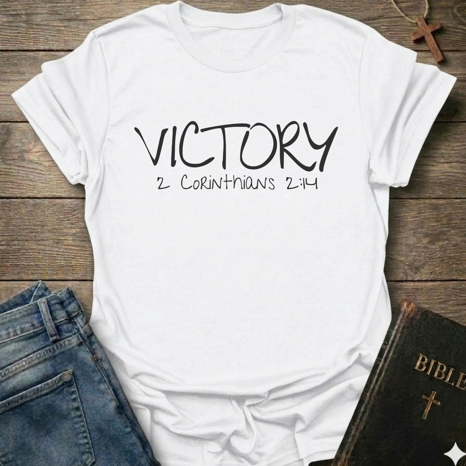 A white blue graphic t-shirt with 'VICTORY 2 CORINTHIANS 2:14' printed in black script, styled in a flat-lay on a wooden background with jeans, a Bible, and a cross necklace.