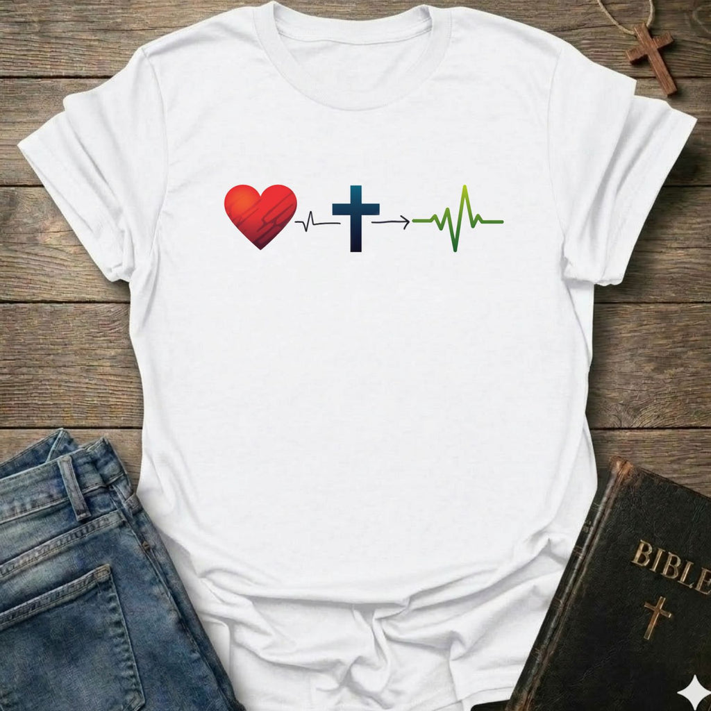 A white short-sleeve T-shirt laid flat on a rustic wooden surface, featuring a graphic of a red heart, a blue cross, and a green heartbeat line. Blue jeans and a black Bible with a gold cross are partially visible as styling props.