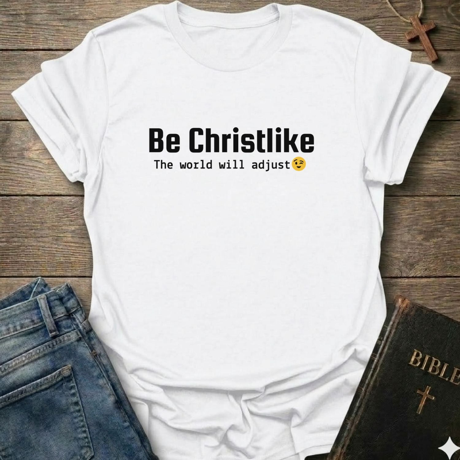 A white short-sleeve graphic T-shirt laid flat on a rustic wooden surface, styled with blue jeans and a Bible. The shirt has black text reading 'Be Christlike' with 'The world will adjust' and a small yellow smiley face underneath.