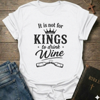 White t-shirt with black text "It is Not for Kings to Drink Wine" and 2 wine bottles graphic based on Proverbs 31:4 Christian faith apparel conversation starter on a wooden surface with jeans and a Bible.