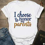 White t-shirt with 'I choose to honor my parents' Exodus 20:12 text navy gold Christian faith apparel conversation starter. T-shirt is on a wooden surface with jeans and a Bible.