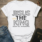 White t-shirt with text 'Jesus The King Over All Kingdoms'  and crown Christian bold faith apparel Faith Forward text on a wooden surface with jeans and a Bible.