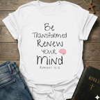 A white graphic t-shirt laid flat on a rustic wooden surface, styled with folded blue jeans and a black Bible. The t-shirt features the text 'Be Transformed Renew Your Mind Romans 12:2' in black lettering with a small red brain icon.
