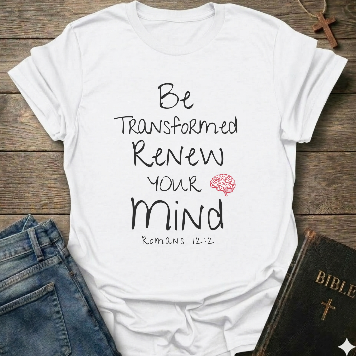 A white graphic t-shirt laid flat on a rustic wooden surface, styled with folded blue jeans and a black Bible. The t-shirt features the text 'Be Transformed Renew Your Mind Romans 12:2' in black lettering with a small red brain icon.