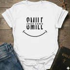 A white short-sleeve graphic t-shirt with a black smiley face design and the text 'SMILE JESUS CARES' and 'SMILL' printed on the front. The shirt is styled in a flat-lay arrangement on a rustic wooden background, accompanied by blue jeans, a brown cross necklace, and a Bible.