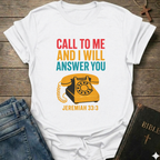 White t-shirt withwith vintage phone graphic and text Call to Me and I Will Answer You Jeremiah 33:3 retro phone faith Christian apparel conversation starter on a wooden surface with jeans and a Bible.