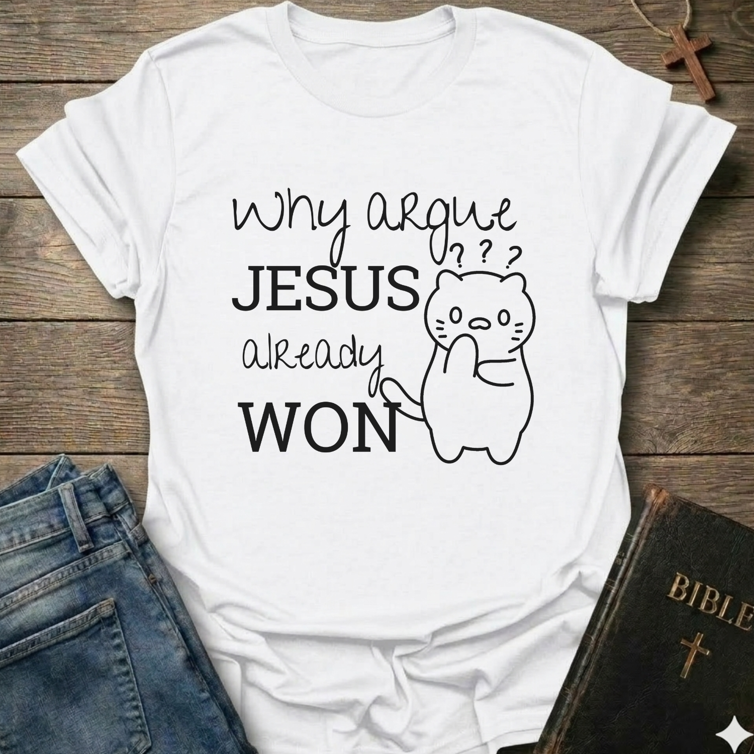 A white graphic t-shirt laid flat on a rustic wooden background, styled with blue jeans and a black Bible. The t-shirt features the text 'Why argue JESUS already WON' in black lettering alongside a simple line drawing of a confused cat.