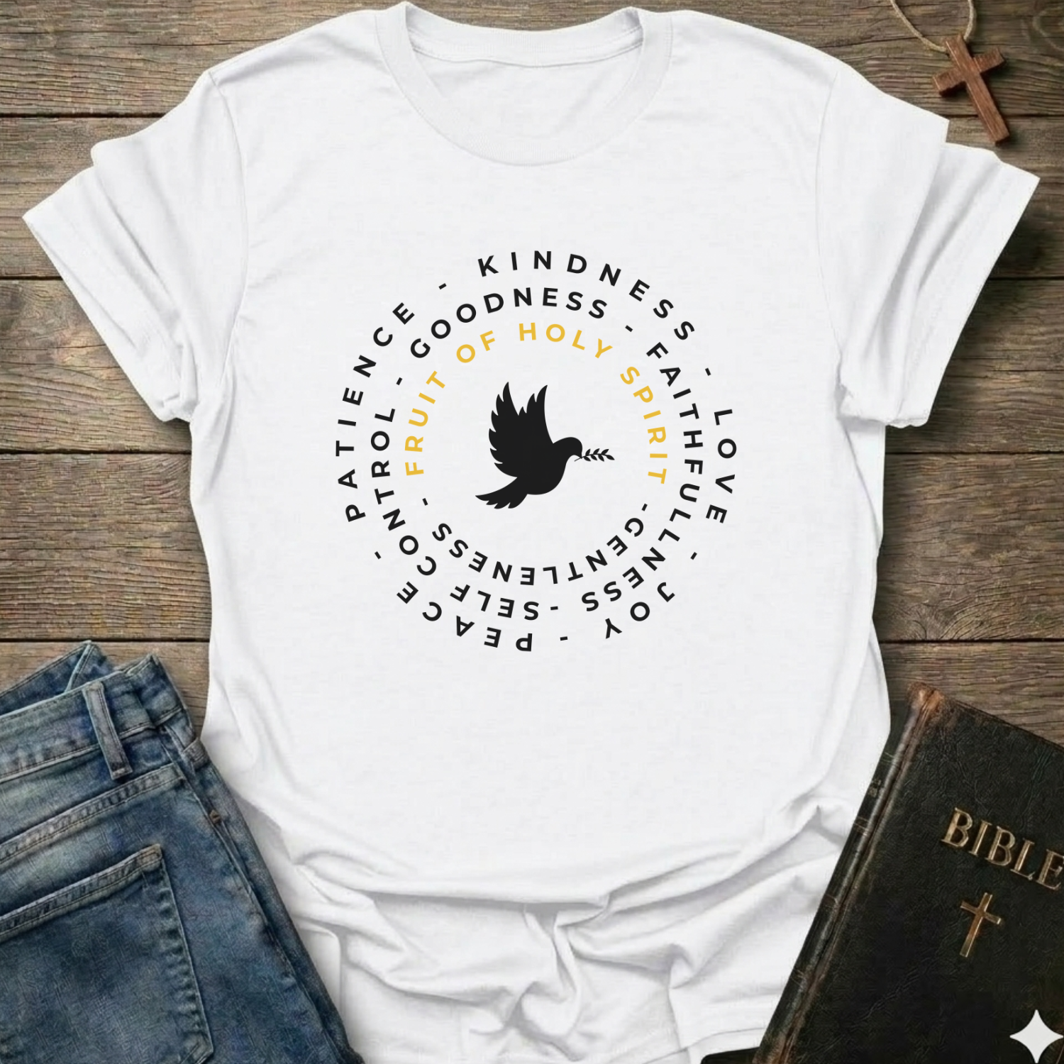 A white short-sleeve T-shirt laid flat on a rustic wooden surface, styled with blue jeans, a Bible, and a wooden cross. The shirt has a circular graphic with a black dove in the center and black text around it, including the words 'PATIENCE', 'KINDNESS', 'GOODNESS', 'FAITH', 'LOVE', 'JOY', 'PEACE', and 'GENTLENESS', with 'FRUIT OF HOLY SPIRIT' in yellow.