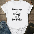A white short-sleeve graphic T-shirt laid flat on a rustic wooden surface, with black text reading 'Mondays are Tough so is My Faith.' Blue jeans and a dark Bible with a cross are partially visible as styling props.