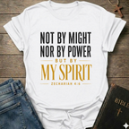 A white graphic t-shirt laid flat on a rustic wooden surface, styled with blue jeans and a black Bible. The t-shirt features the text 'NOT BY MIGHT NOR BY POWER BUT BY MY SPIRIT ZECHARIAH 4:6' in black and gold lettering.