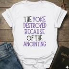White t-shirt with text 'The yoke destroyed because of the anointing' Isaiah 10:27 faith apparel conversation starter on a wooden surface with jeans and a Bible.