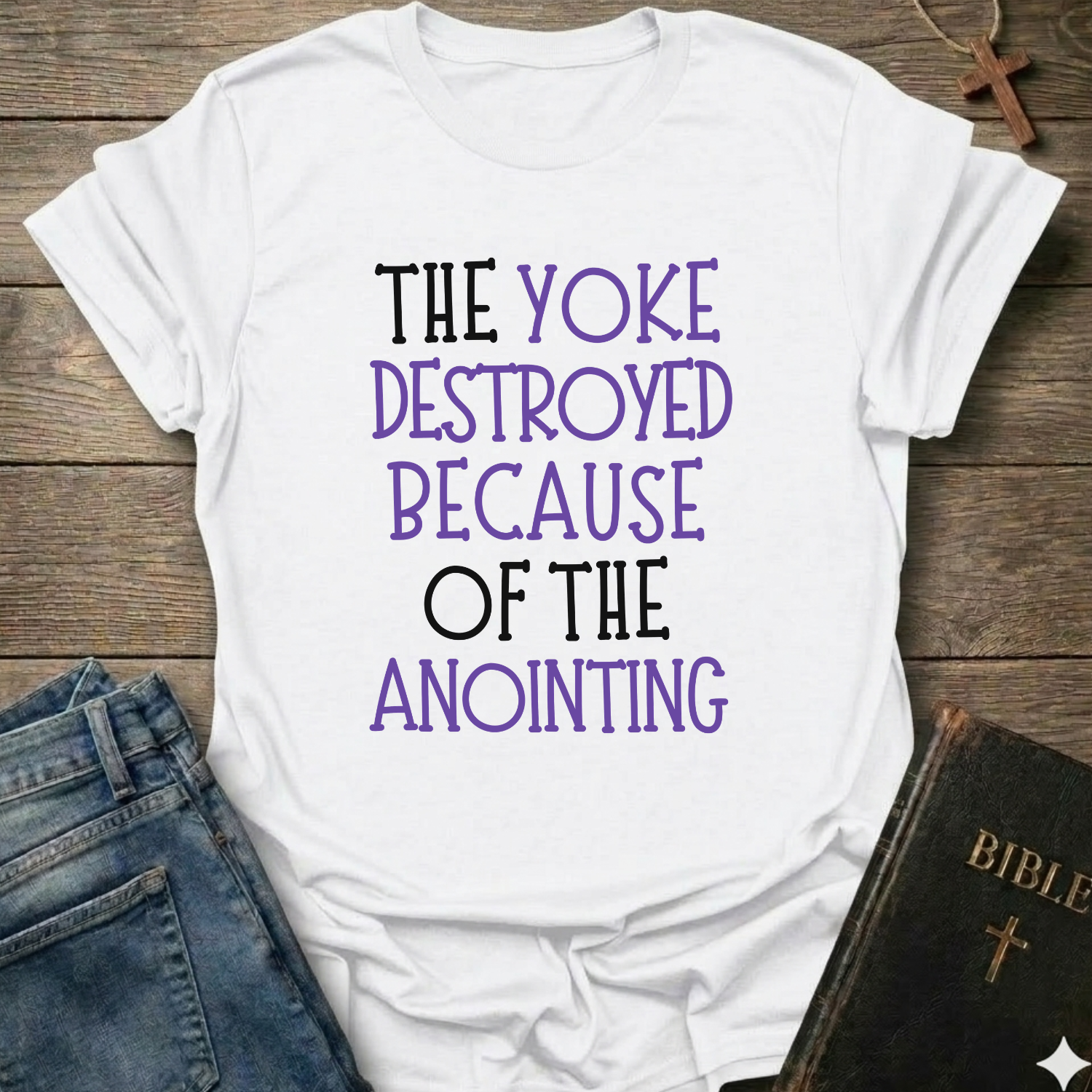 White t-shirt with text 'The yoke destroyed because of the anointing' Isaiah 10:27 faith apparel conversation starter on a wooden surface with jeans and a Bible.