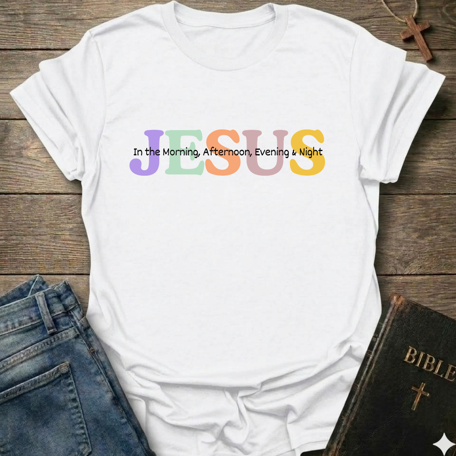 A white short-sleeve T-shirt laid flat on a rustic wooden background, styled with blue jeans, a Bible, and a cross necklace. The shirt features the word 'JESUS' in large, colorful pastel letters (purple, green, orange, yellow) with the phrase 'In the Morning, Afternoon, Evening & Night' printed in black script on top of it.