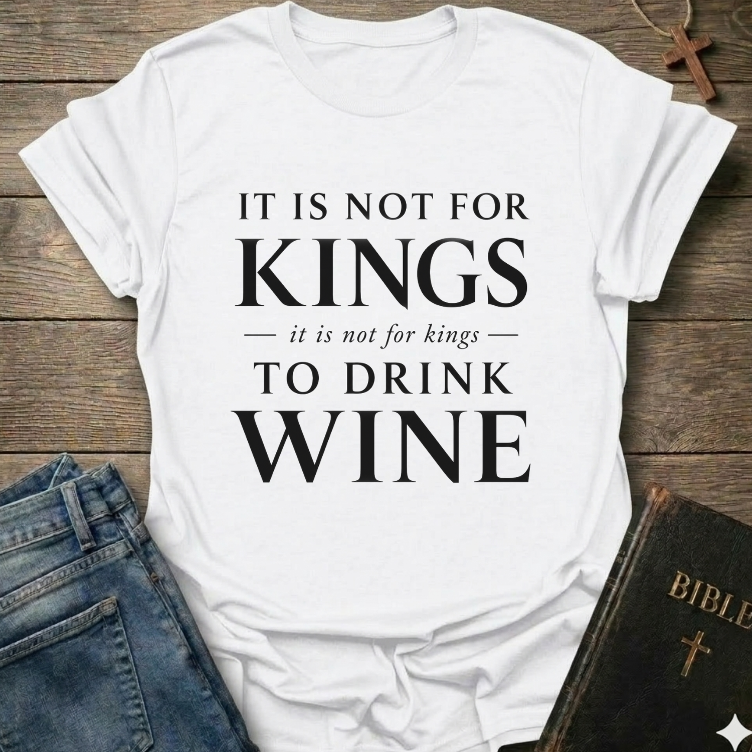 A white short-sleeve graphic T-shirt laid flat on a rustic wooden surface, with black text reading 'IT IS NOT FOR KINGS it is not for kings TO DRINK WINE.' Blue jeans and a dark Bible are partially visible as styling props.