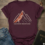 Maroon Christian t-shirt with 'Faith can move mountains' graphic Matthew 17:20 faith apparel conversation starter on a wooden surface with jeans and a Bible. 