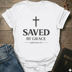 White Christian t-shirt with 'Saved by Grace' text and cross reference Ephesians 2:8 scripture faith apparel conversation starter  on a wooden surface with jeans and a Bible. 