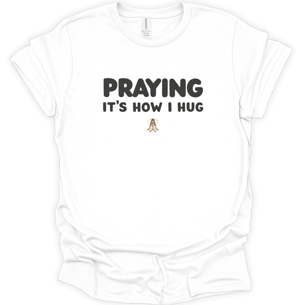 A white short-sleeve crewneck t-shirt with the text 'PRAYING IT'S HOW I HUG' printed in black on the chest, with a small praying figure graphic below.
