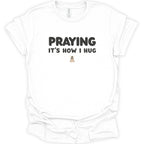 A white short-sleeve crewneck t-shirt with the text 'PRAYING IT'S HOW I HUG' printed in black on the chest, with a small praying figure graphic below.