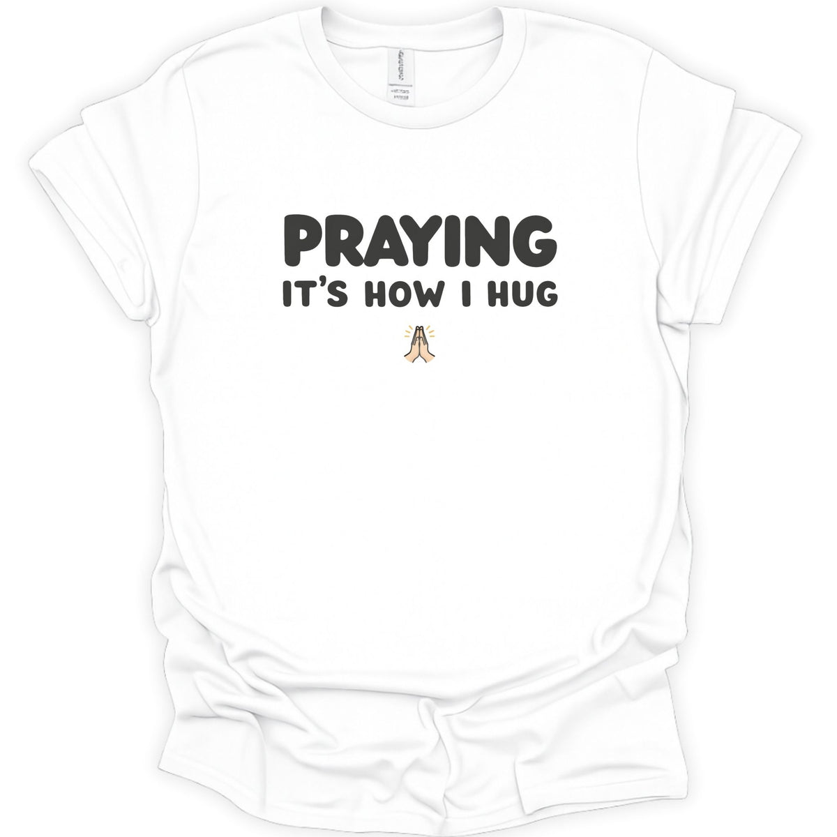 A white short-sleeve crewneck t-shirt with the text 'PRAYING IT'S HOW I HUG' printed in black on the chest, with a small praying figure graphic below.