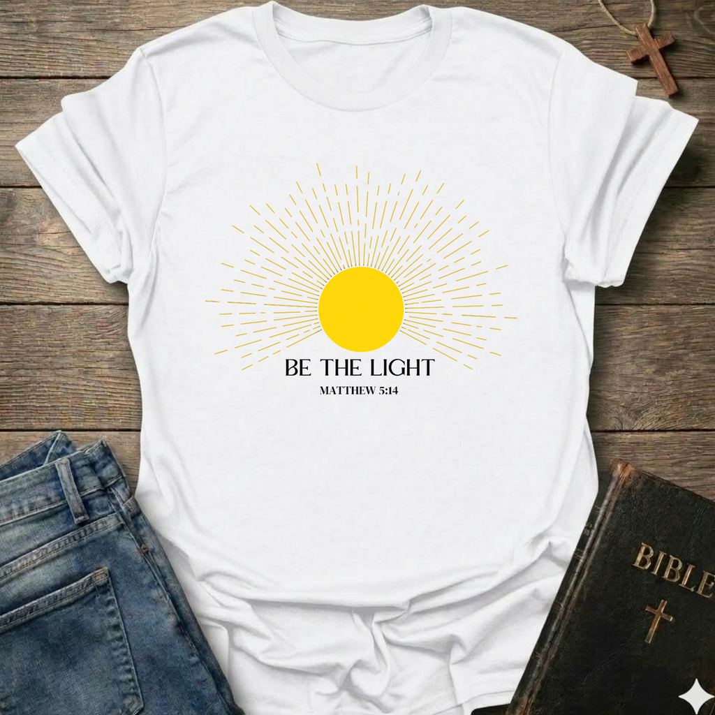 White t-shirt with yellow sun graphic and 'Be the Light' text on a wooden surface with jeans and a Bible. Be the Light Christian T-Shirt Matthew 5:14 bold faith apparel Faith Forward