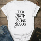 White t-shirt with 'Seek Truth Find Jesus' text on a wooden surface with jeans and a Bible. Bold typography faith apparel conversation starter.