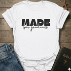 A white short-sleeve T-shirt laid flat on a rustic wooden surface, with the text 'MADE for greatness' based on 2 Corinthians 5 verse 17 printed on the front, styled in a flat-lay arrangement with blue jeans and a Bible on a rustic wooden background.