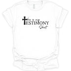 A white short-sleeve crewneck t-shirt with a black graphic text print reading 'this is my TESTIMONY Shirt' accompanied by a cross icon, displayed on a clean white background.