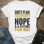 A white graphic t-shirt laid flat on a rustic wooden background, styled with blue jeans and a Bible. The t-shirt features the text 'GOD'S PLAN: PROSPERITY, HOPE & A FUTURE FOR ME!' in black and mustard yellow lettering.