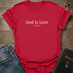 A red short-sleeve crewneck t-shirt with the text 'God is Love 1 John 4:16' printed in white on the chest. The shirt is styled in a flat-lay arrangement on a rustic wooden background, accompanied by blue jeans and a black leather-bound Bible.