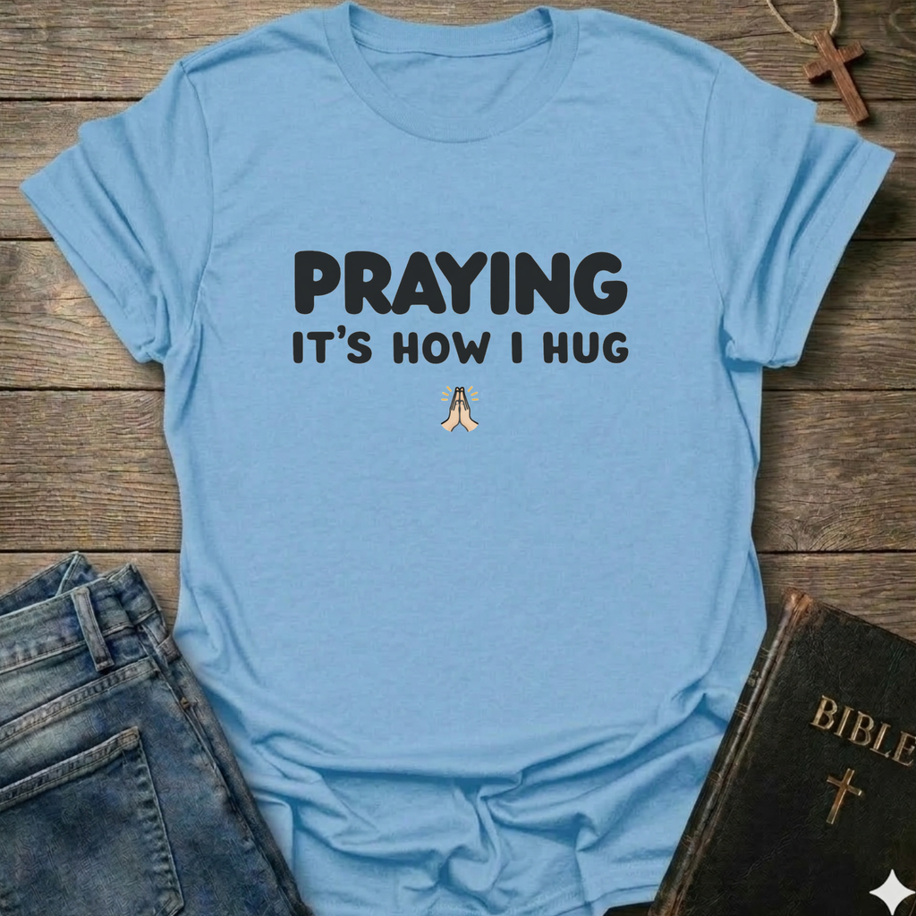 Light blue t-shirt with 'PRAYING IT'S HOW I HUG' text Christian faith apparel relatable scripture tee conversation starter on a wooden surface with jeans and a Bible.