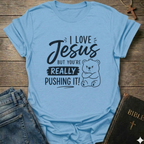 A light blue graphic t-shirt laid flat on a rustic wooden background, styled with a pair of blue jeans on the left and a black Bible with a gold cross on the right. The t-shirt features a black printed graphic with the text 'I LOVE Jesus BUT YOU'RE REALLY PUSHING IT!' and a cartoon bear illustration.