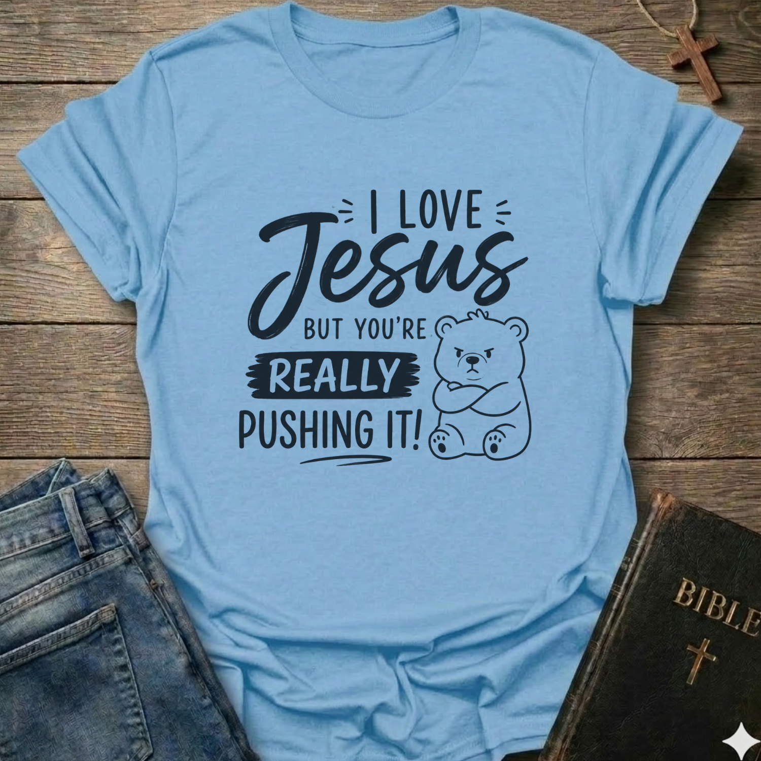 A light blue graphic t-shirt laid flat on a rustic wooden background, styled with a pair of blue jeans on the left and a black Bible with a gold cross on the right. The t-shirt features a black printed graphic with the text 'I LOVE Jesus BUT YOU'RE REALLY PUSHING IT!' and a cartoon bear illustration.