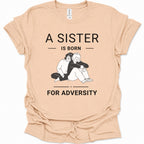 A natural-colored short-sleeve t-shirt with a black graphic print that reads 'A SISTER IS BORN FOR ADVERTS' surrounding an illustration of two people hugging. The shirt is laid flat on a white background.