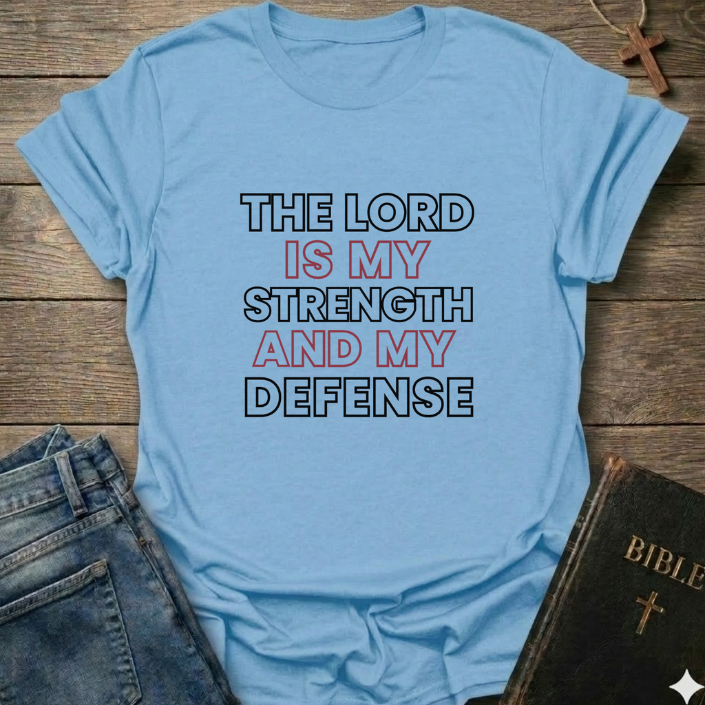 Light blue t-shirt with 'The Lord is my strength and my defense' Exodus 15:2 bold faith apparel Faith Forward on a wooden surface with jeans and a Bible.