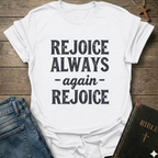 White t-shirt with 'REJOICE ALWAYS again REJOICE' text Philippians 4:4 bold faith apparel Faith Forward on a wooden surface with jeans and a Bible.