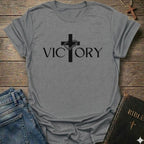 A sport gray short-sleeve t-shirt with a black graphic print of a cross and the word 'VICTORY' laid flat on a rustic wooden surface, styled with blue jeans and a Bible.
