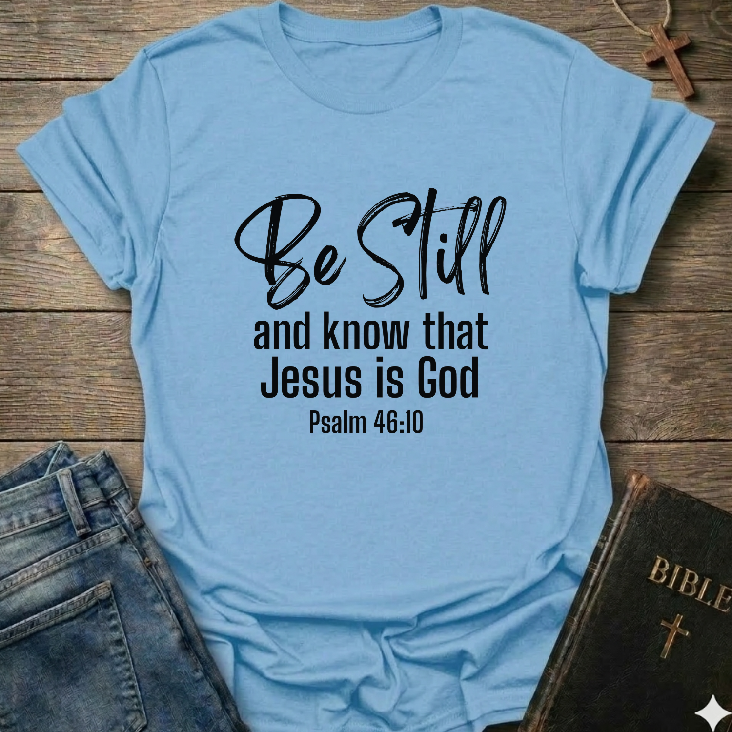 Light blue t-shirt with 'Be Still and know that Jesus is God' text Psalm 46:10 script faith apparel Faith Forward on a wooden surface with jeans and a Bible.