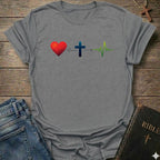 A sport gray short-sleeve t-shirt laid flat on a rustic wooden surface, featuring a graphic of a red heart connected to a blue Christian cross by a heartbeat line, which leads to a green heartbeat line. Blue jeans and a black Bible with a gold cross are styled as props around the shirt.