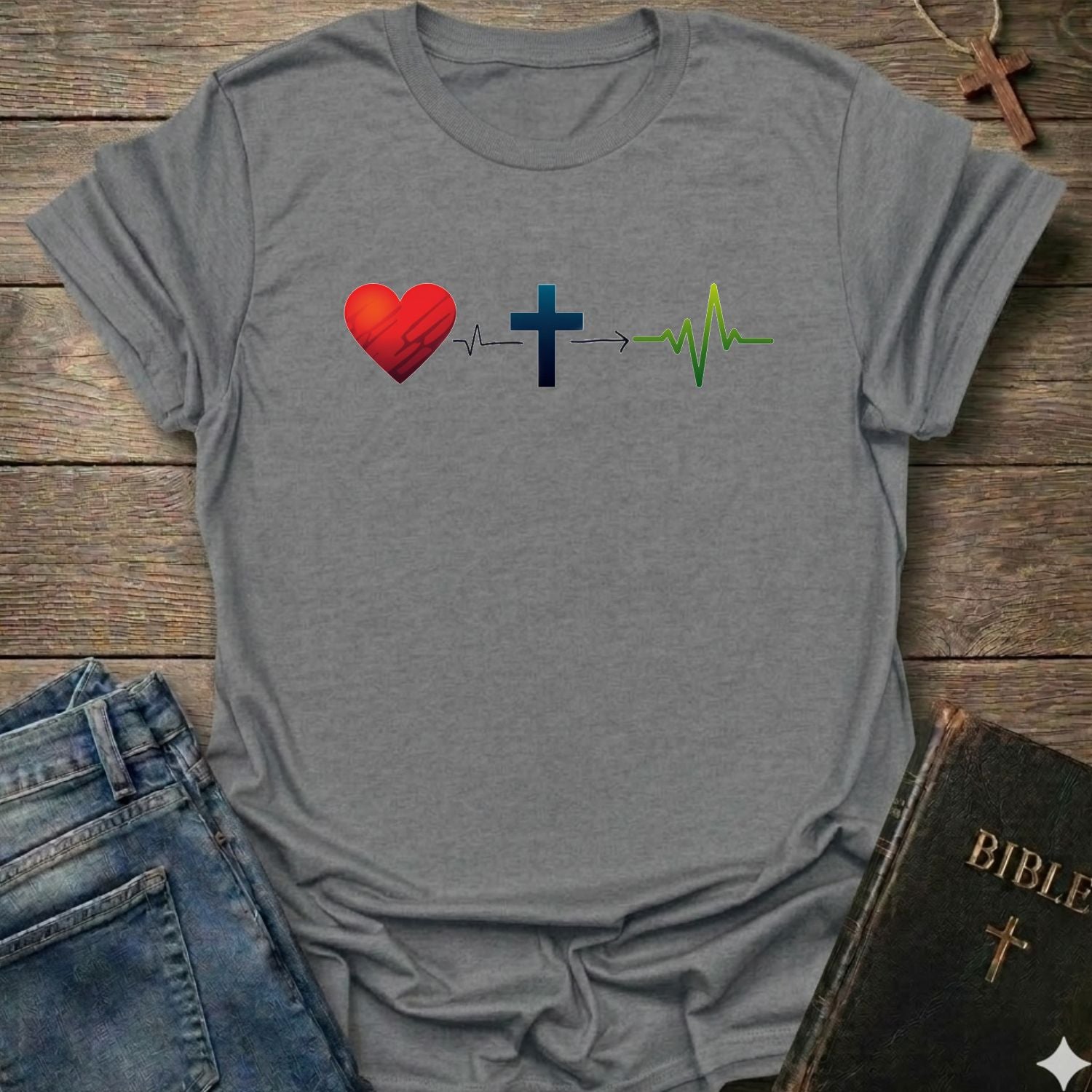 A sport gray short-sleeve t-shirt laid flat on a rustic wooden surface, featuring a graphic of a red heart connected to a blue Christian cross by a heartbeat line, which leads to a green heartbeat line. Blue jeans and a black Bible with a gold cross are styled as props around the shirt.