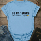Light blue t-shirt with 'Be Christlike The world will adjust' text on a wooden surface with jeans and a Bible.
