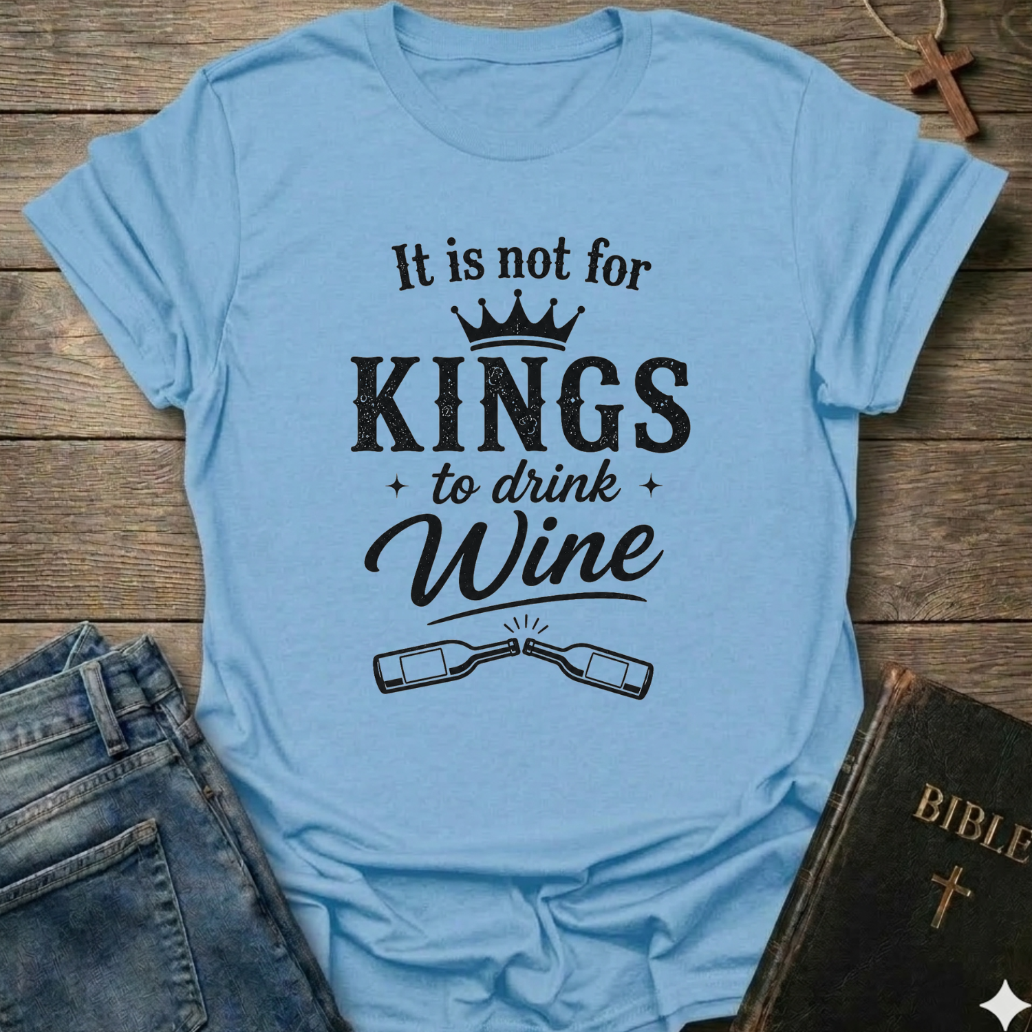Light blue t-shirt with black text "It is Not for Kings to Drink Wine" and 2 wine bottles graphic based on Proverbs 31:4 Christian faith apparel conversation starter on a wooden surface with jeans and a Bible.