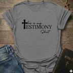 Gray t-shirt with 'This is my testimony shirt' text and cross minimal faith apparel conversation starter. It is on a wooden surface with jeans and a Bible.