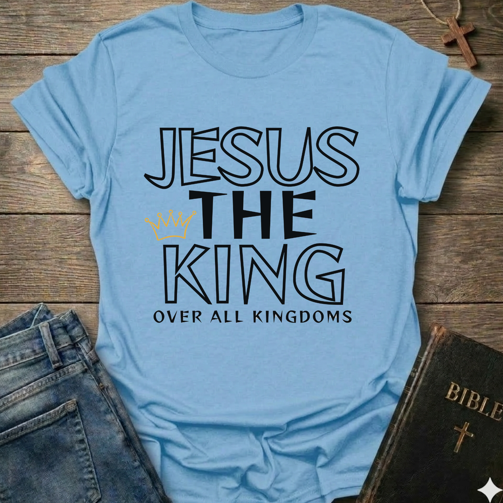 Light blue t-shirt with text 'Jesus The King Over All Kingdoms'  and crown Christian bold faith apparel Faith Forward text on a wooden surface with jeans and a Bible.