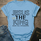 Light blue t-shirt with text 'Jesus The King Over All Kingdoms'  and crown Christian bold faith apparel Faith Forward text on a wooden surface with jeans and a Bible.
