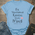 A light blue graphic t-shirt laid flat on a rustic wooden background, styled with blue jeans and a Bible. The t-shirt features the text 'Be Transformed Renew Your Mind Romans 12:2' in black lettering with a small red brain illustration.
